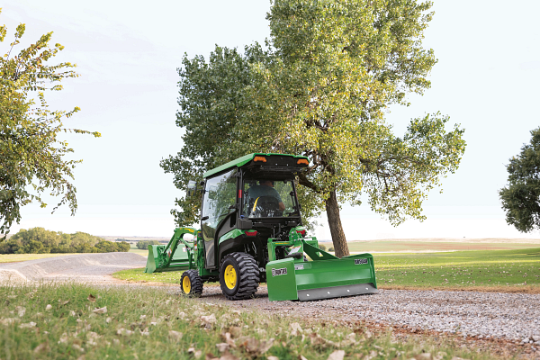 A green tractor with an enclosed cab pulls a box blade attachment along a gravel path beside a tree, with grassy fields and a flat rural landscape under a clear sky