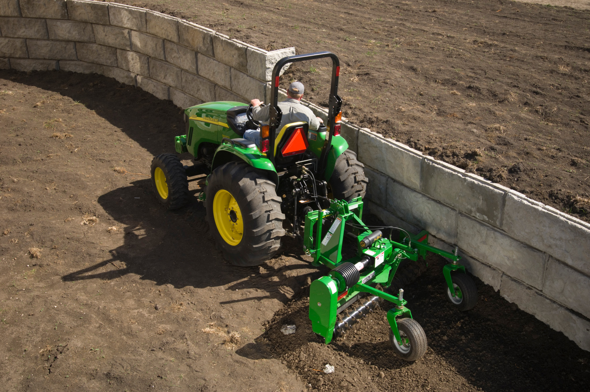 Frontier power rake hitched to a small tractor raking dirt along a stone retaining wall