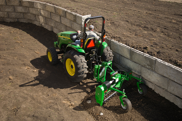 Frontier power rake hitched to a small tractor raking dirt along a stone retaining wall