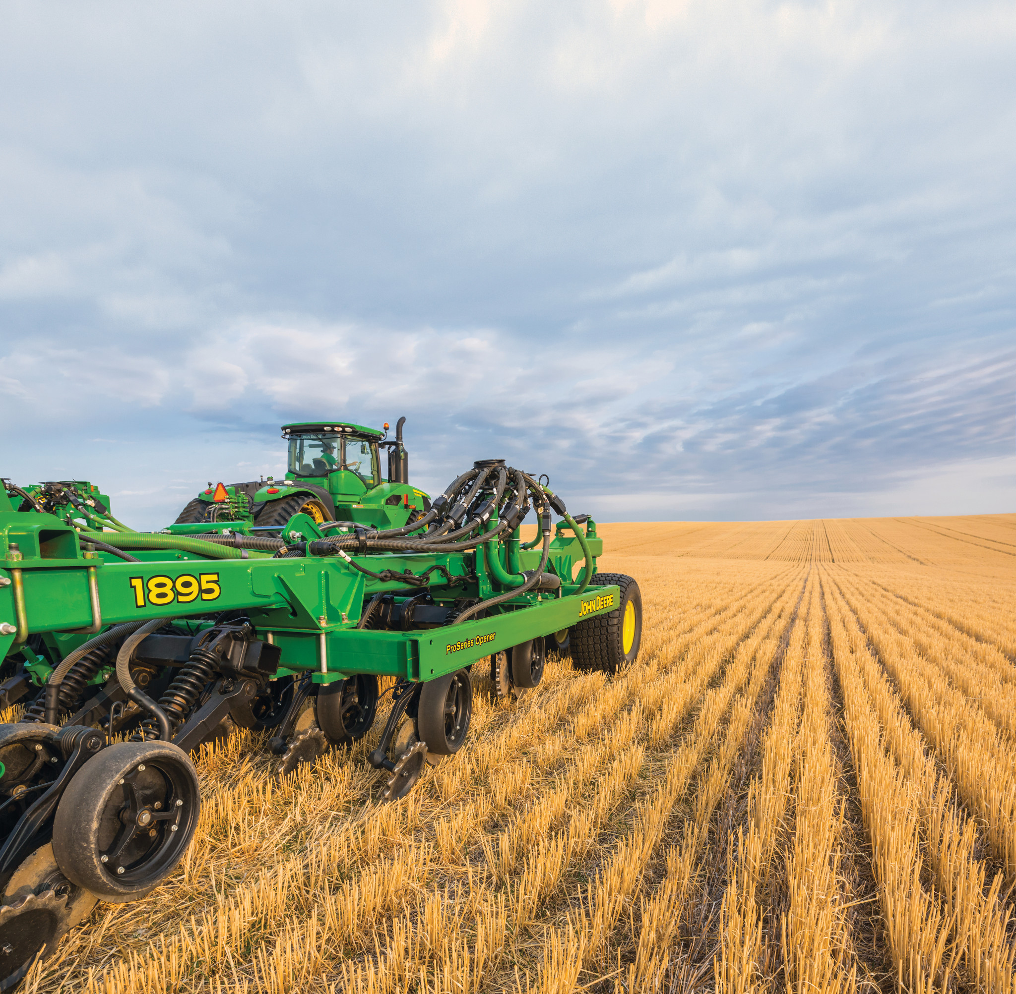 Large green agricultural tractor pulling a wide tillage implement across a harvested field of golden corn stubble, with long parallel rows stretching into the distance under a dramatic, overcast sky