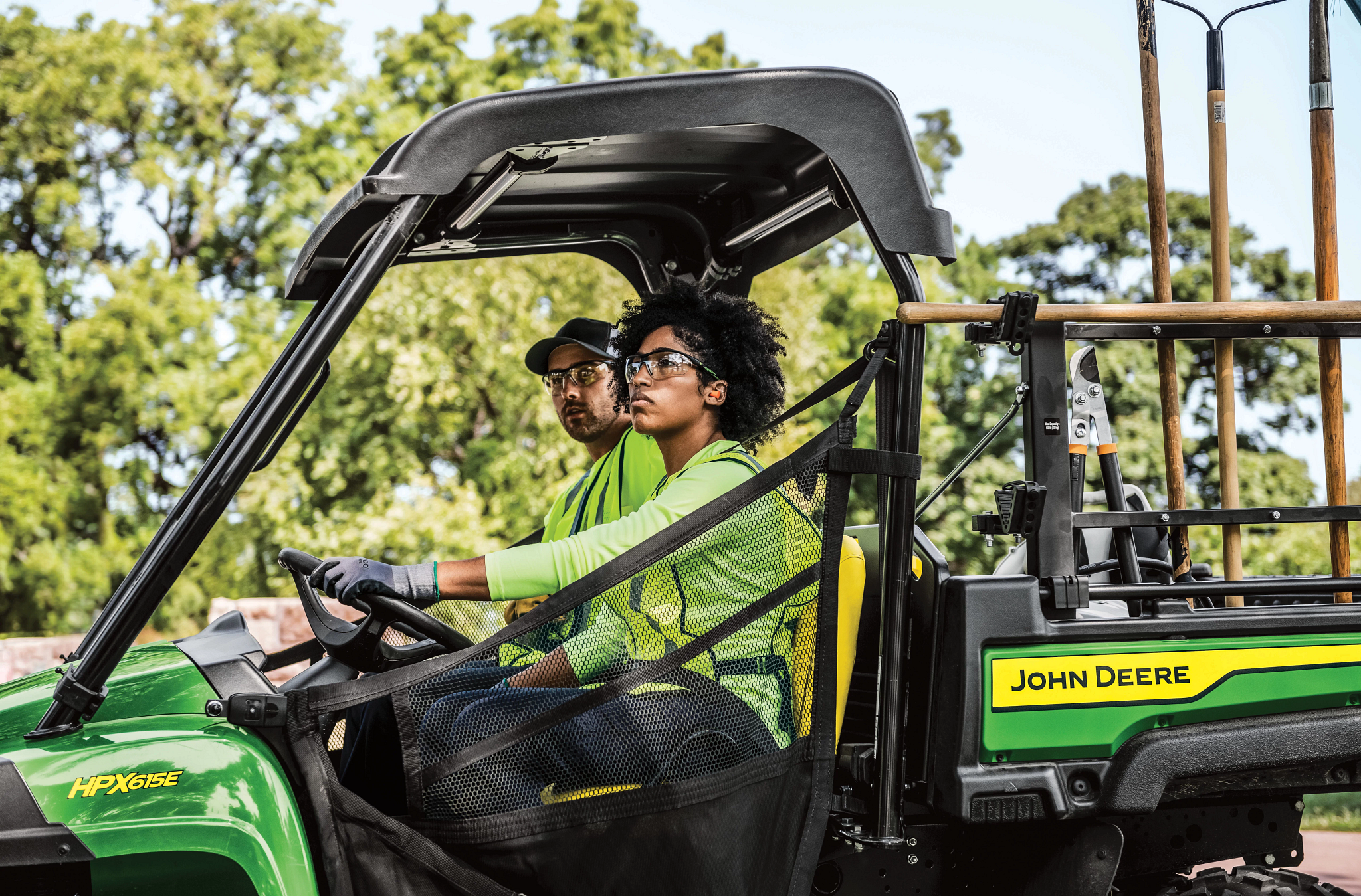 Side left view of two landscapers in an HPX615E Work Utility Vehicle with rakes, shears, and other tools visible in the cargo box