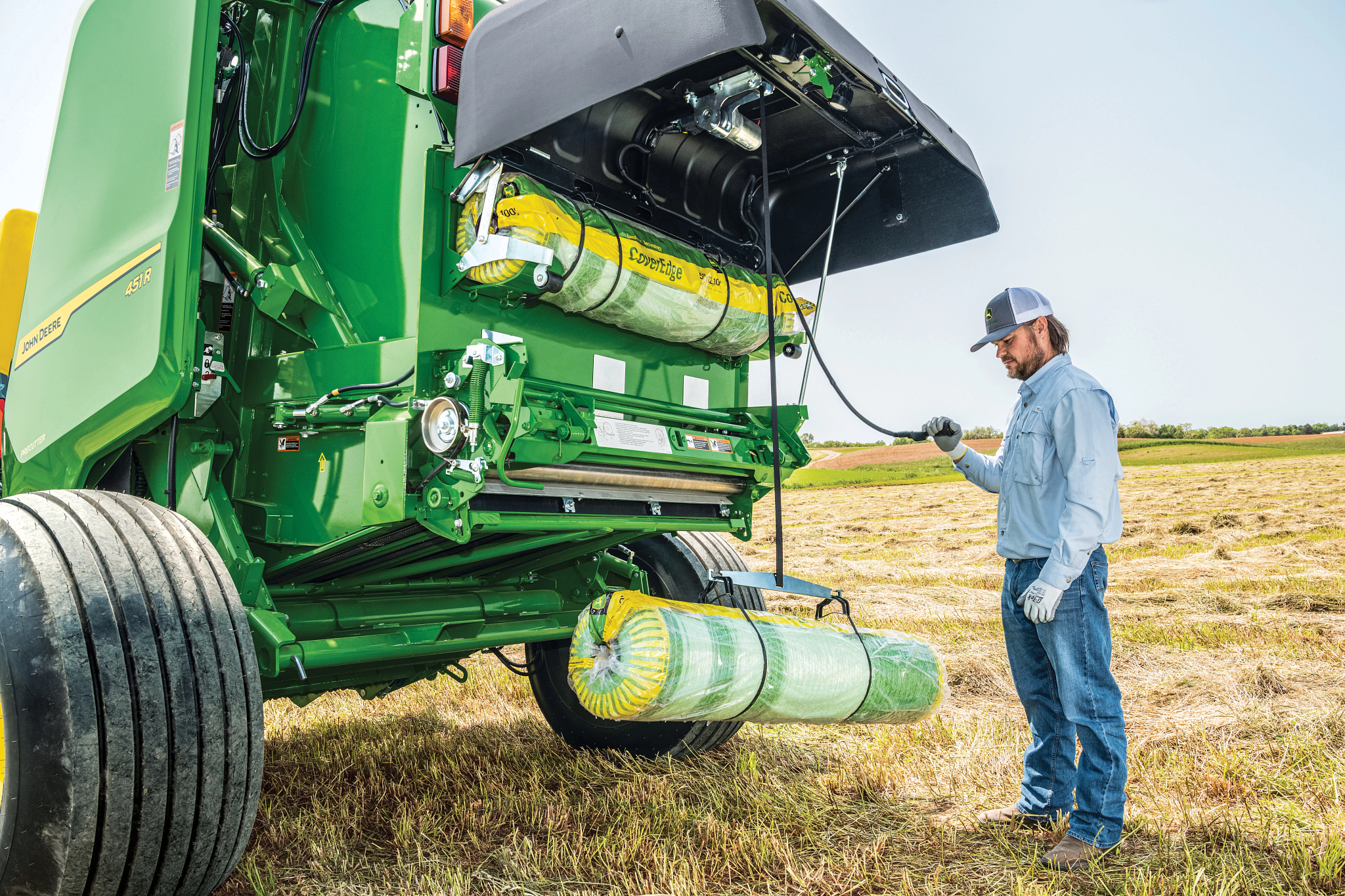 John Deere round baler with rear gate open in a harvested field as an operator inspects the machine.