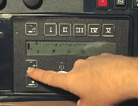 A close-up of a control panel with rectangular buttons, while a hand presses a lower left button beside a display screen showing symbols and settings