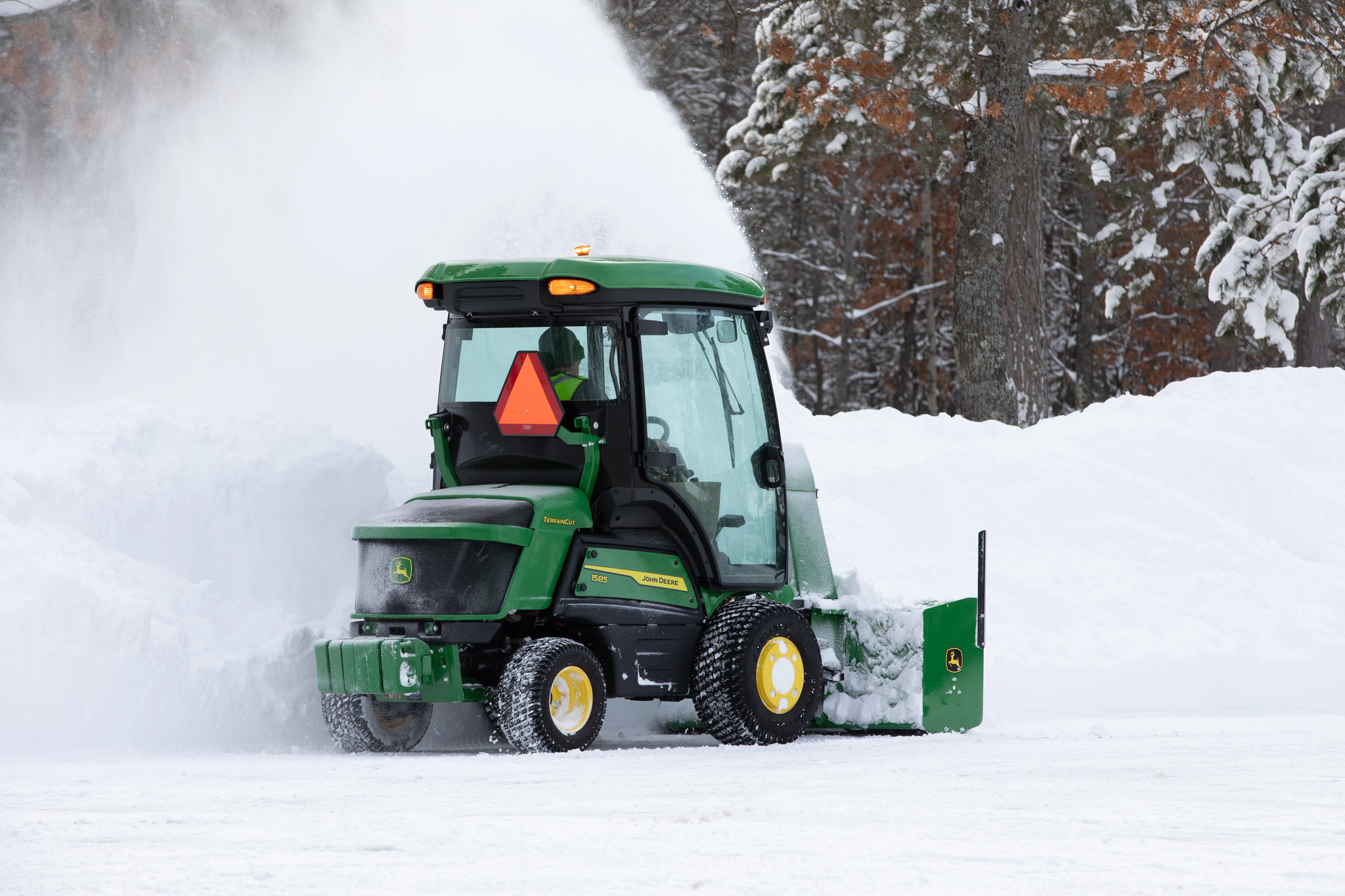 Rear right view of a 1585 TerrainCut™ mower with cab and snowblower attached throwing snow on a tree-lined road