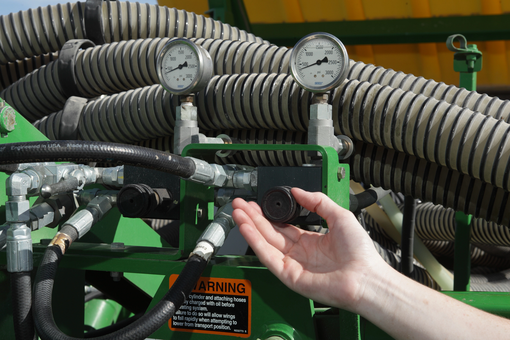 A person’s hand reaches toward a hydraulic component on a nutrient applicator, with hoses, metal fittings, and two pressure gauges visible