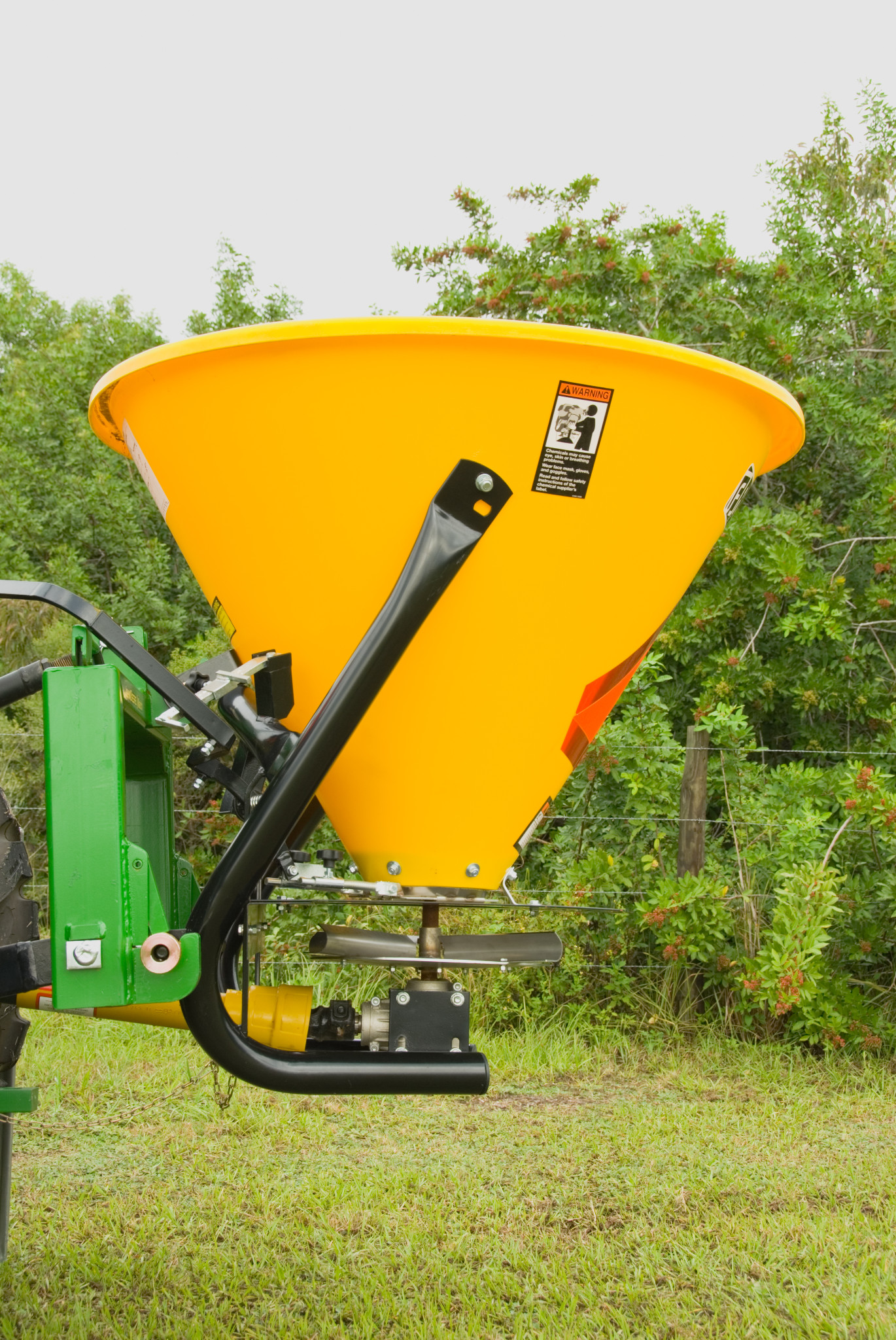 Yellow cone-shaped broadcast spreader mounted to a green frame, shown outdoors with grass and trees in the background