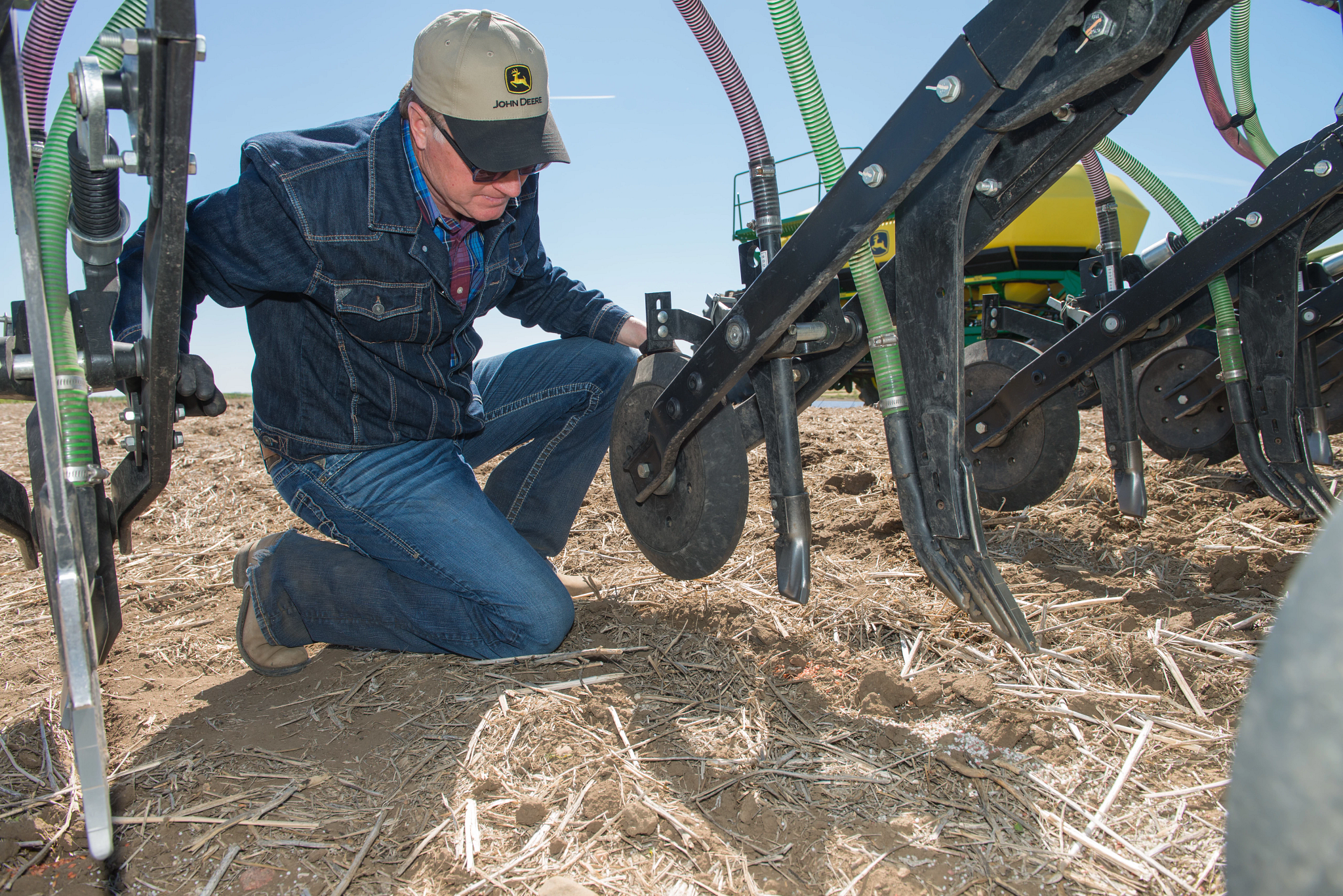 Man kneeling on a field beside an air hoe drill, inspecting the ground-engaging components with metal discs and hoses arranged in a row on dry soil under a clear sky