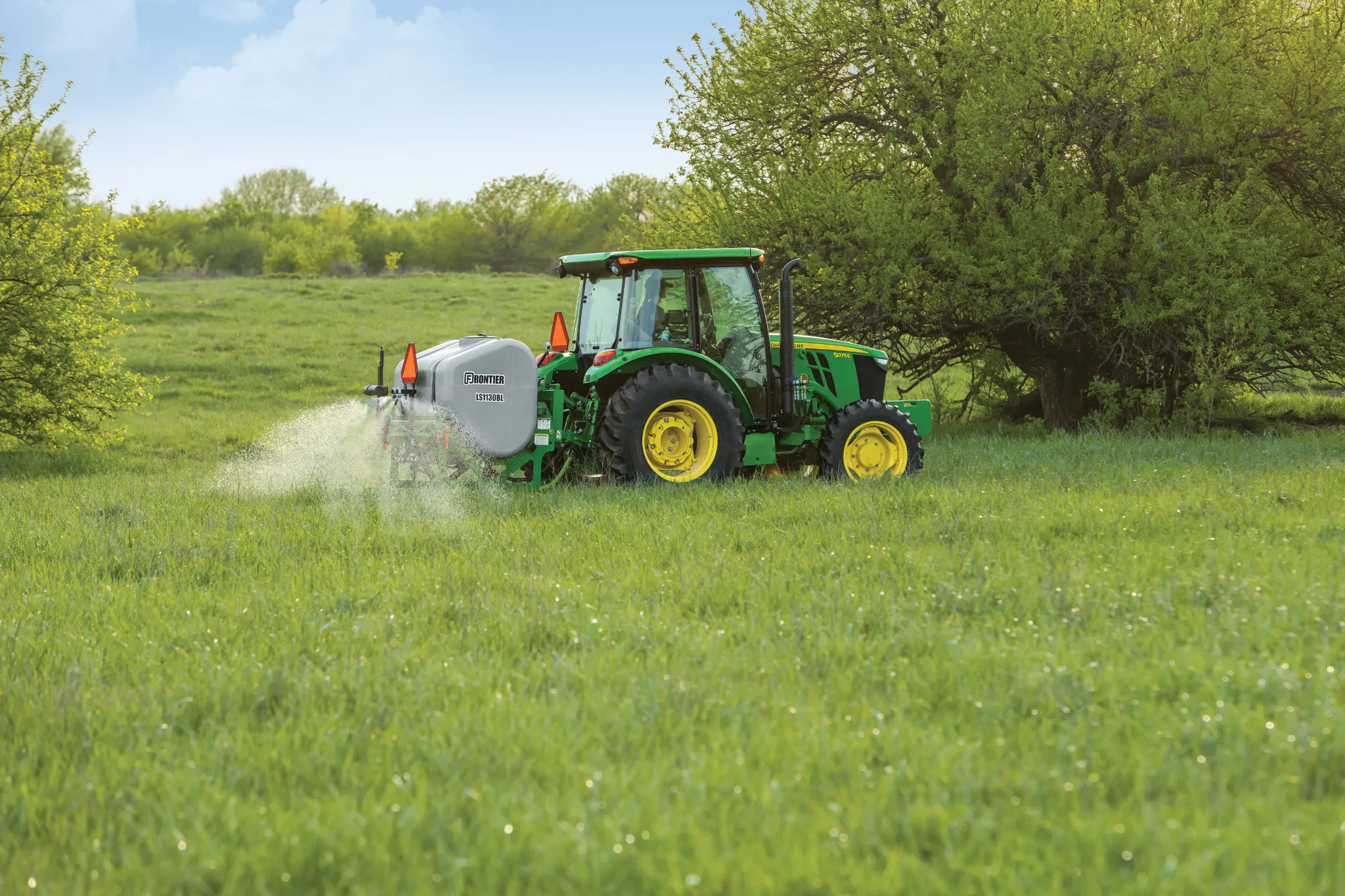 A tractor pulls a sprayer across a lush, grassy field, releasing a fine mist near a tree line on a sunny day.