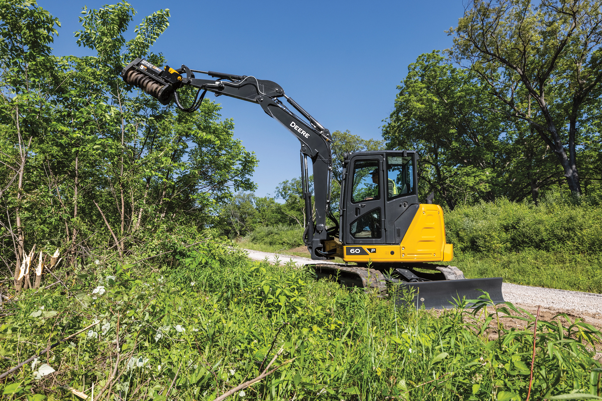 Compact yellow John Deere excavator with a black arm fitted with a brush-cutting attachment, clearing dense green vegetation along a narrow gravel path