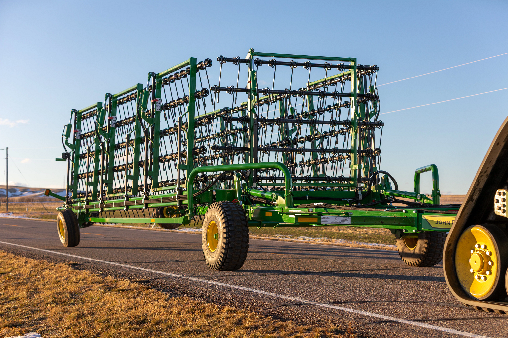 Front right view of a folded HH70 Heavy Harrow being transported on a road between fields. The corner of a 9RX Tractor's rear track is seen in the shot's right side.