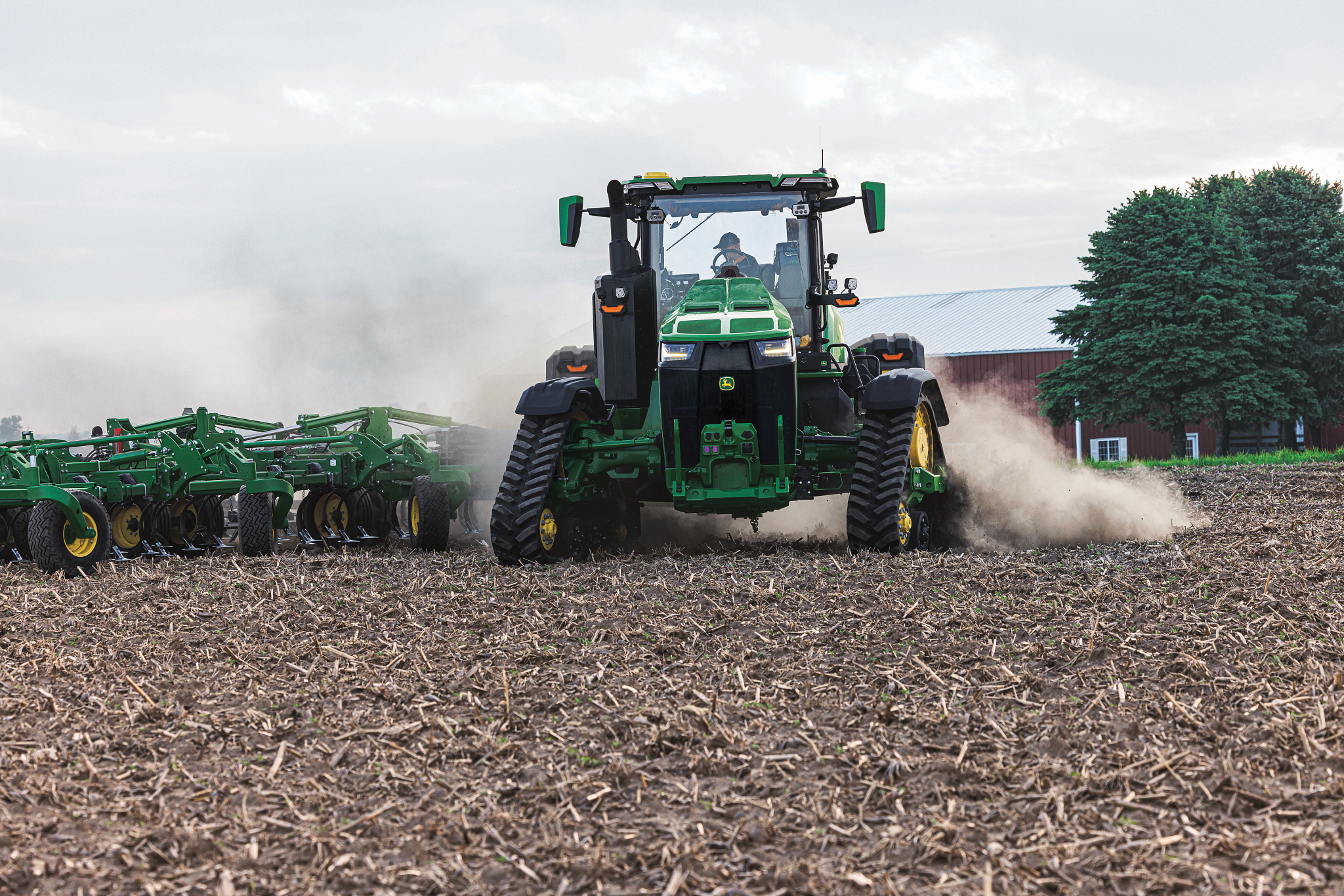 Front view of a large green tractor pulling tillage equipment across a harvested field covered in crop residue with dust rising from the machine and trees and a cloudy sky visible in the background