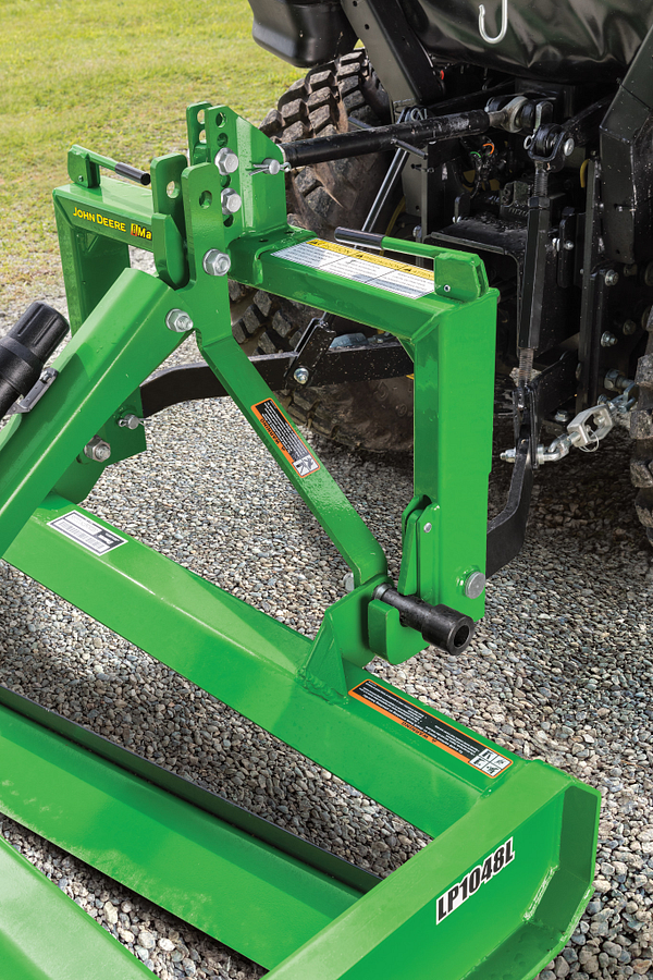 John Deere LP1048L pallet fork attachment with green steel frame and fork tines, mounted on a tractor using a quick‑attach system, shown outdoors on gravel with tractor tire and linkage visible.