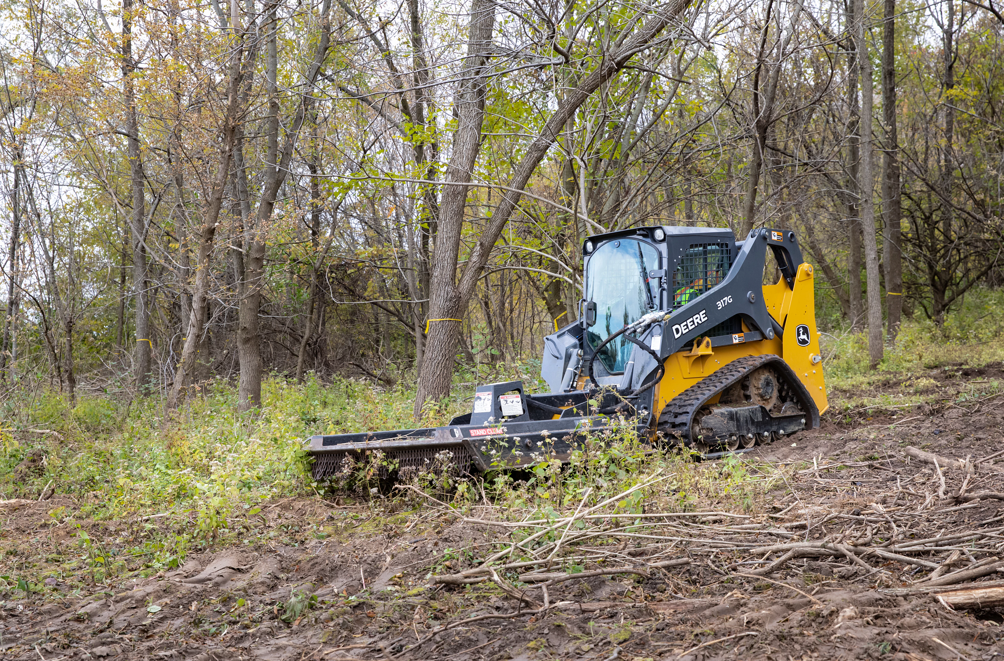 Front left view of a 317G compact track loader in the woods with front-attached RC72B rotary cutter, clearing shrubs and sticks from a muddy clearing