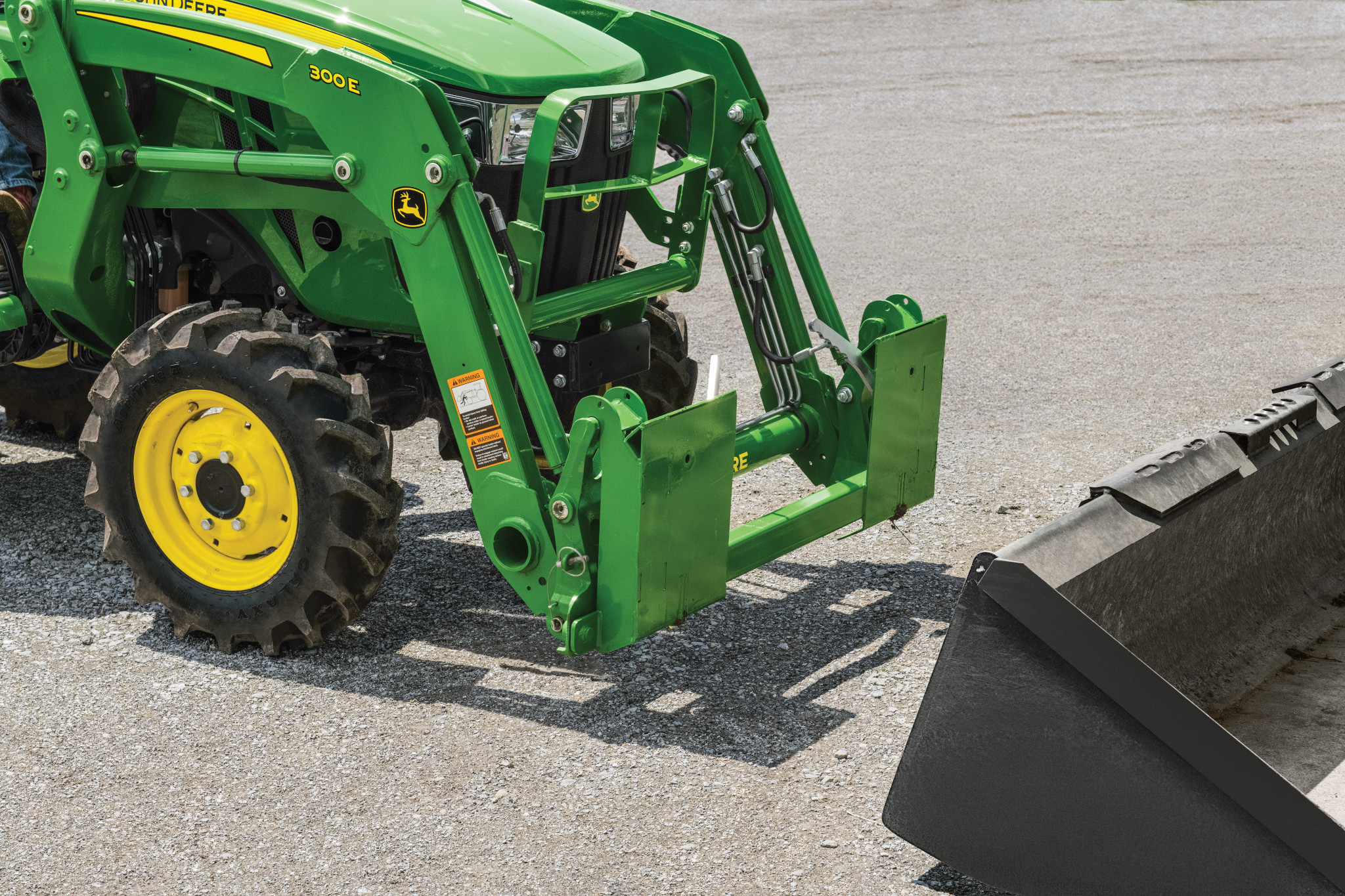 Front loader of a green tractor positioned in front of a detached bucket, showing the carrier adapter and hydraulic connections on a paved surface