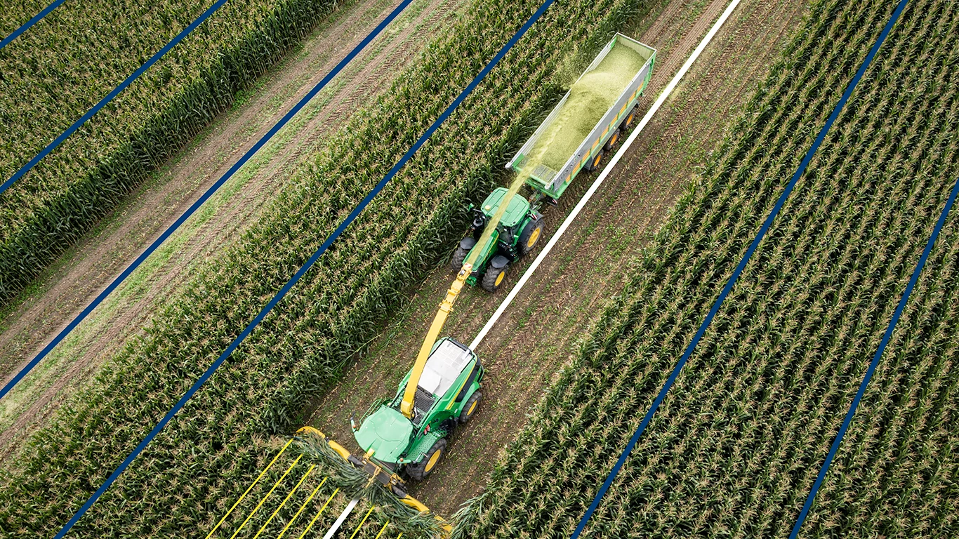 John Deere forage harvester transferring chopped crop into a trailer while driving between corn rows, viewed from above with guidance lines