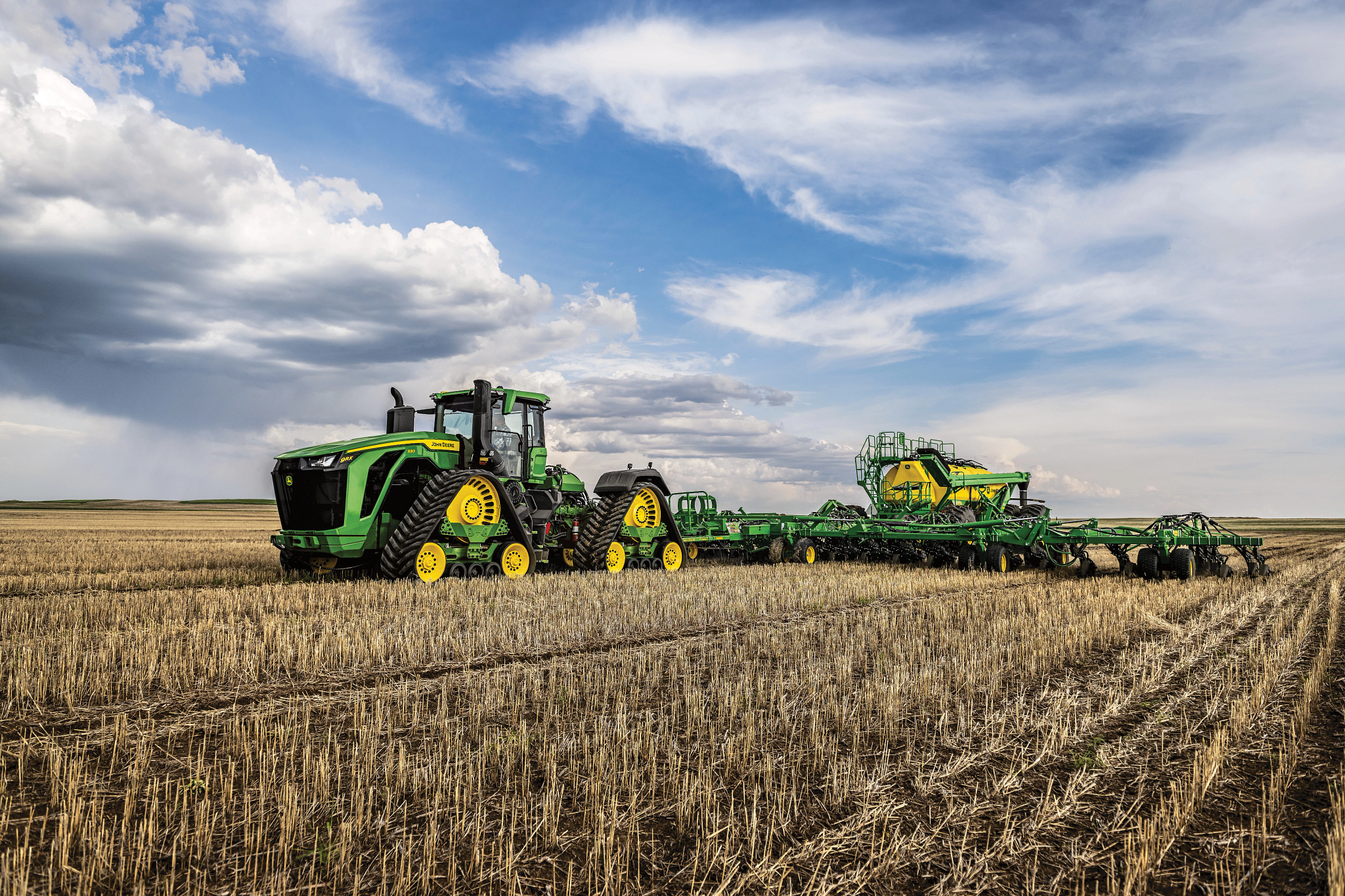 Green tractor with dual tracks pulling a long green seeding implement across a harvested field of stubble under a dramatic sky with scattered clouds stretching over open farmland