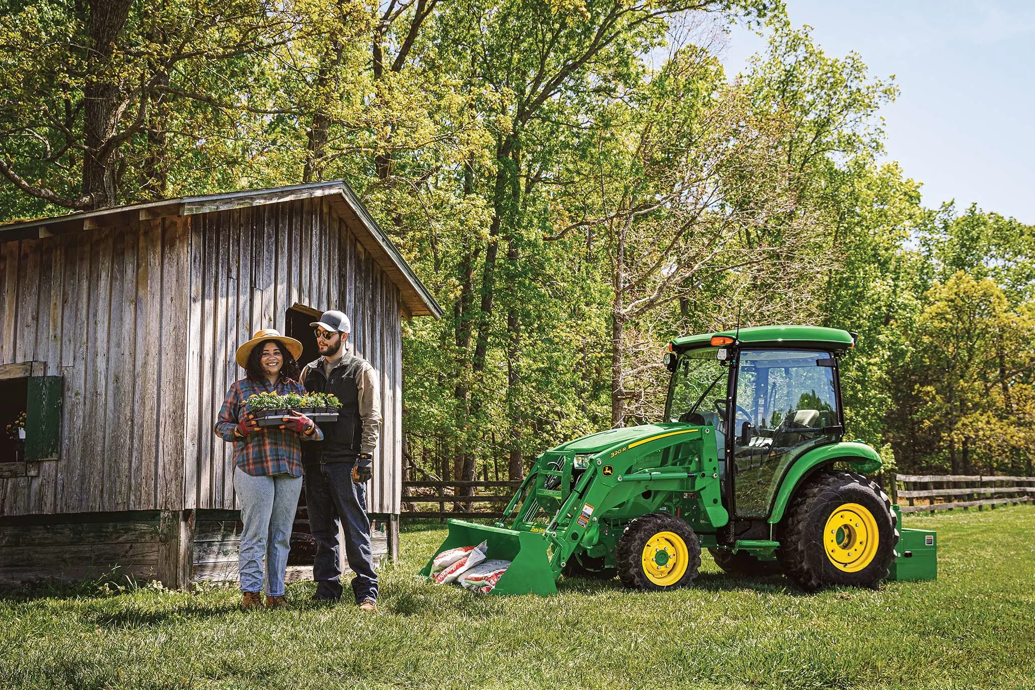 Two farmers standing in front of a wood building in front of a fenced-off field and woods, with a green 3046R compact utility tractor parked to the right of them.