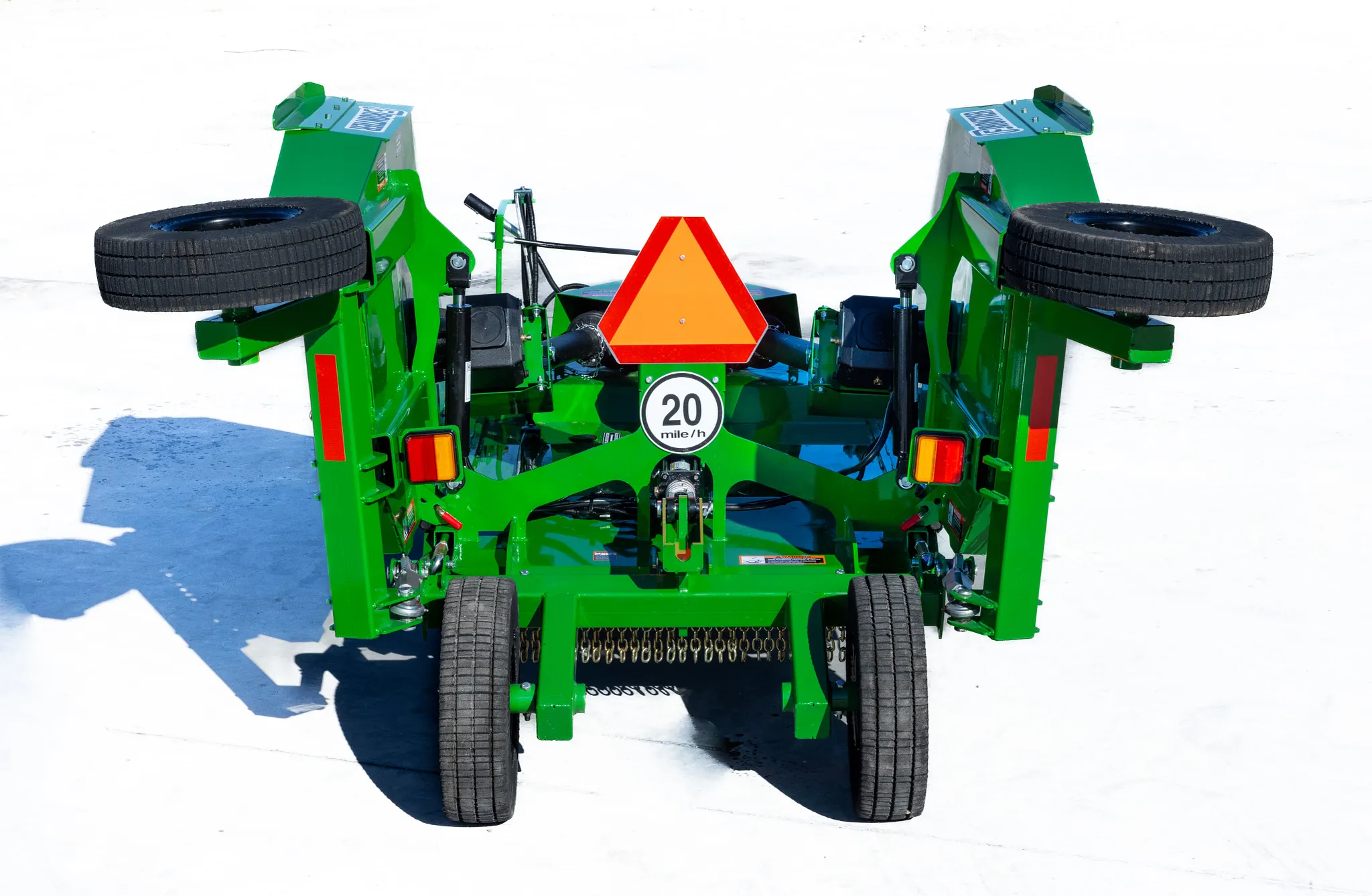 Rear view of a green Frontier rotary cutter in transport position, showing raised wing decks, transport wheels, lights, safety chain shielding, a 20 mph sign, and a slow-moving vehicle emblem on a concrete surface