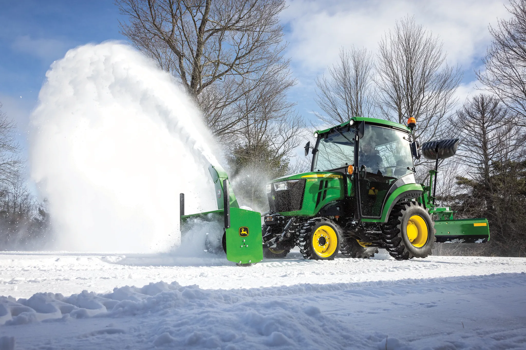 Green tractor equipped with a front-mounted snow blower clearing a snowy path, blasting a high arc of snow to the side next to bare trees and under a cloudy sky