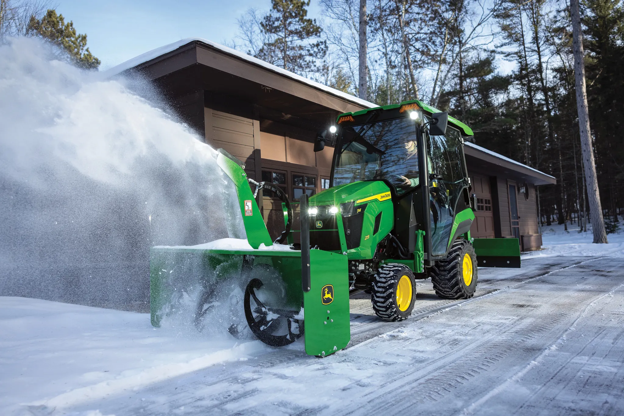 A green tractor equipped with a front-mounted snow blower clears snow from a driveway in front of a garage, blowing a wide spray of snow to the side among tall trees
