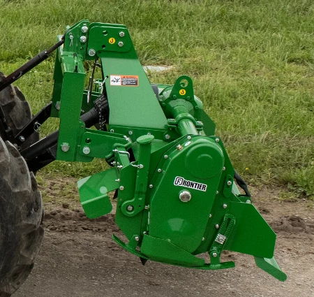 Side left view of a green-painted Frontier rotary tiller hovering over dirt and grass from the back of a tractor. The edges of the tractor's back tires are visible.