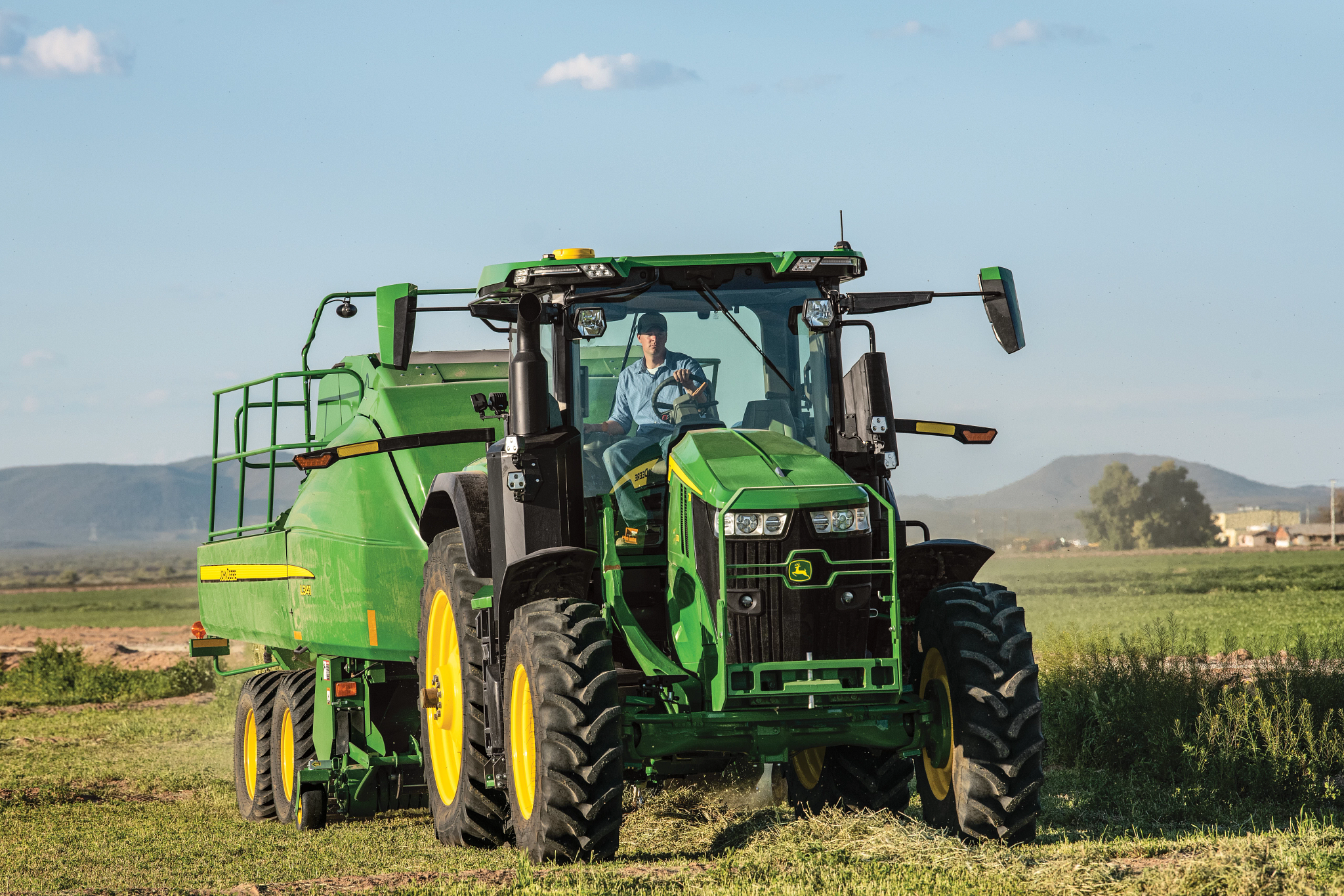 Green tractor pulling a large baler across a grassy farm field, with wide tires and a glass cab, set against a flat rural landscape with distant hills and a clear blue sky