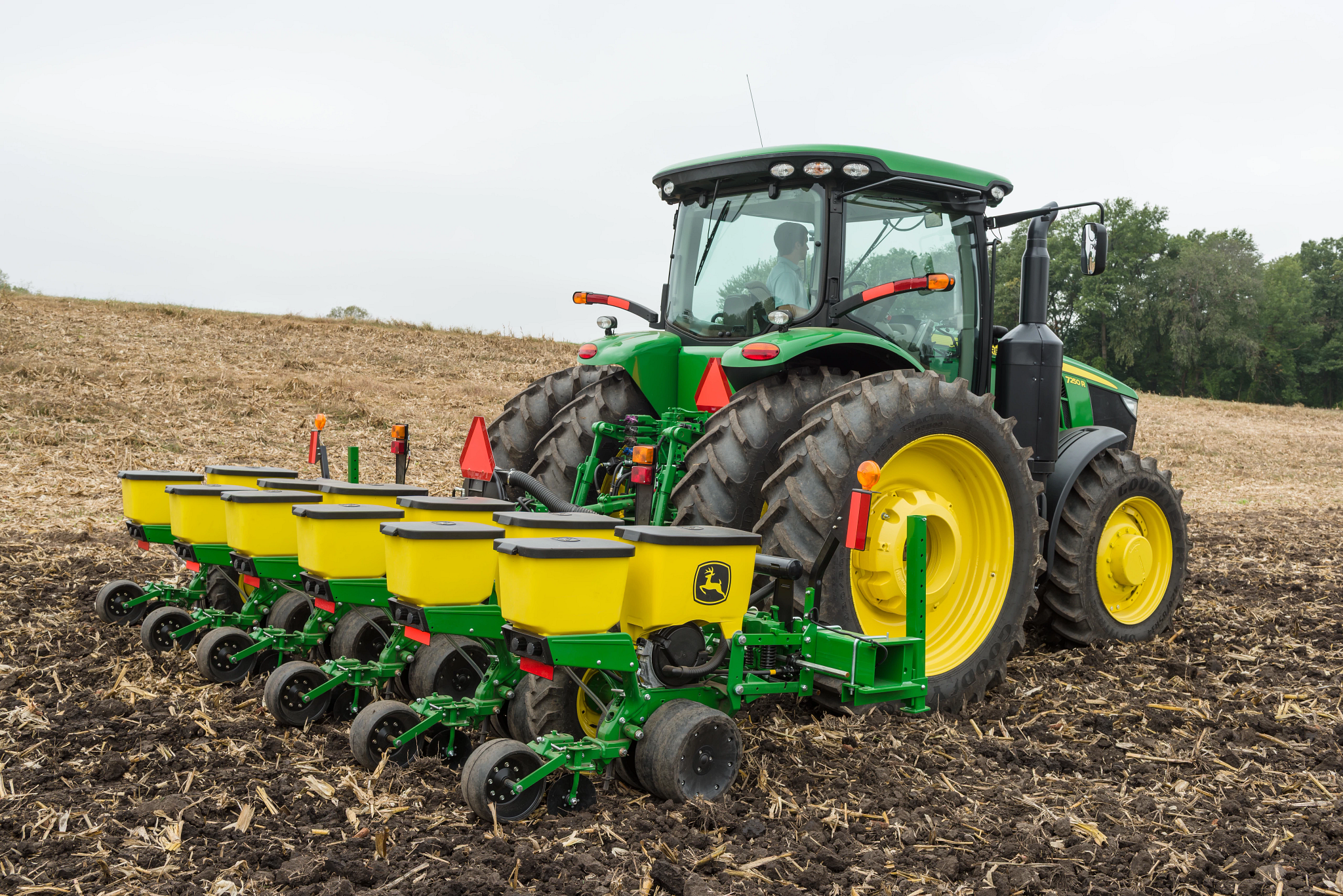 Tractor with large yellow wheels pulling a multi-row planter across a brown, freshly tilled field, with crop residue visible on the soil and a line of trees in the background under an overcast sky.