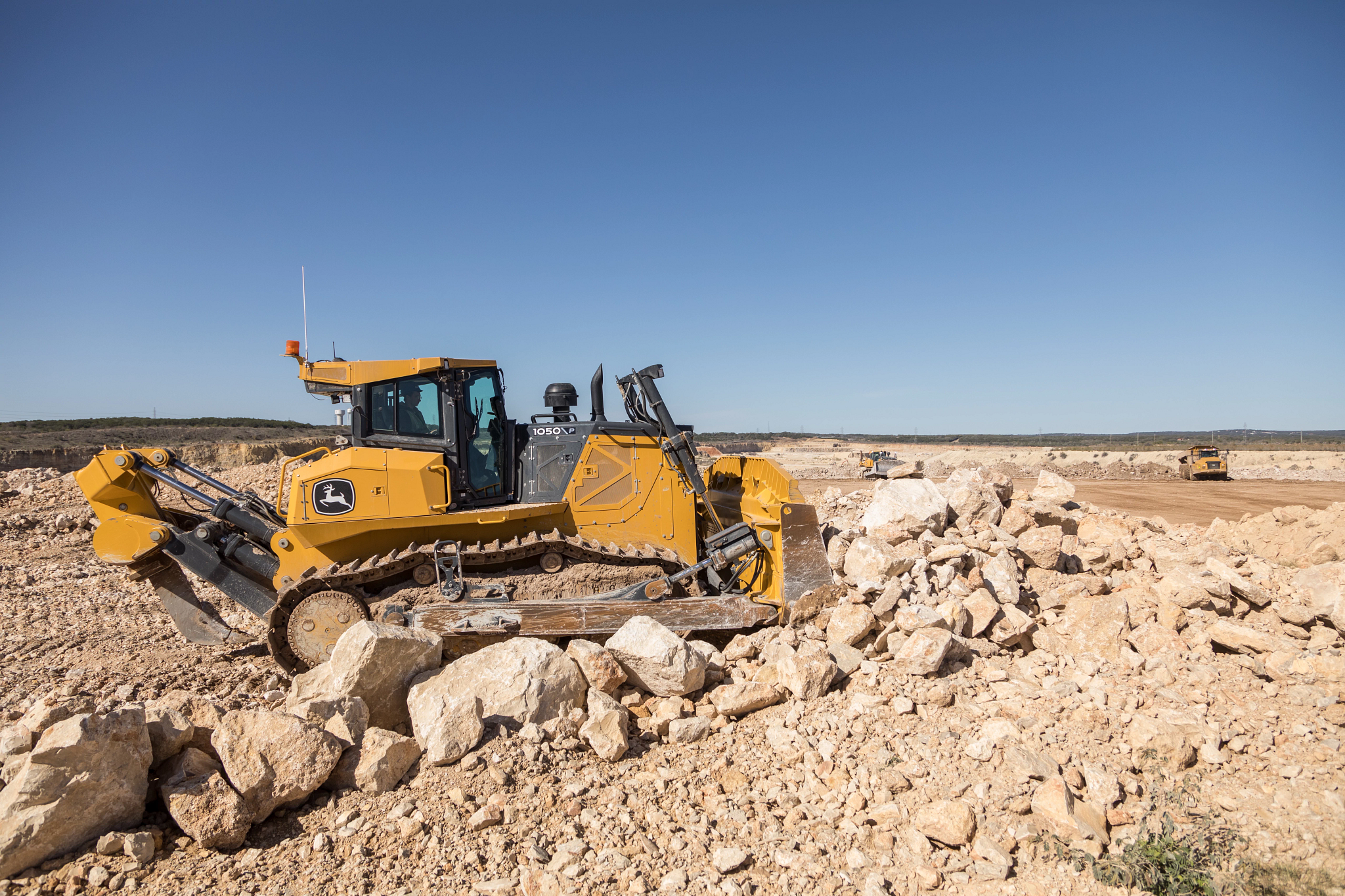 John Deere skid steer loader with a wide front grading attachment leveling soil at a construction site, with a commercial building, dirt piles, and clear blue sky in the background.