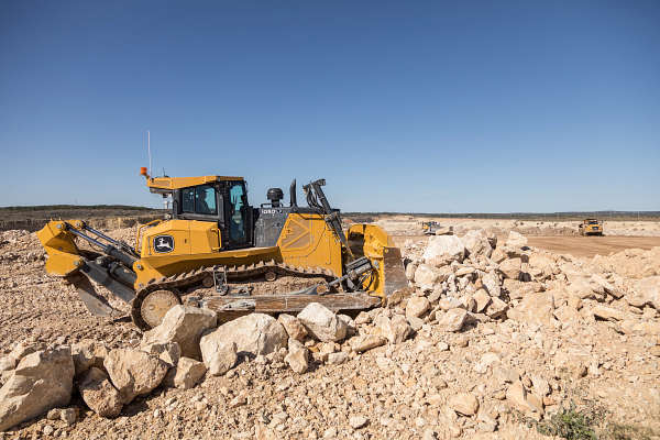 John Deere skid steer loader with a wide front grading attachment leveling soil at a construction site, with a commercial building, dirt piles, and clear blue sky in the background.