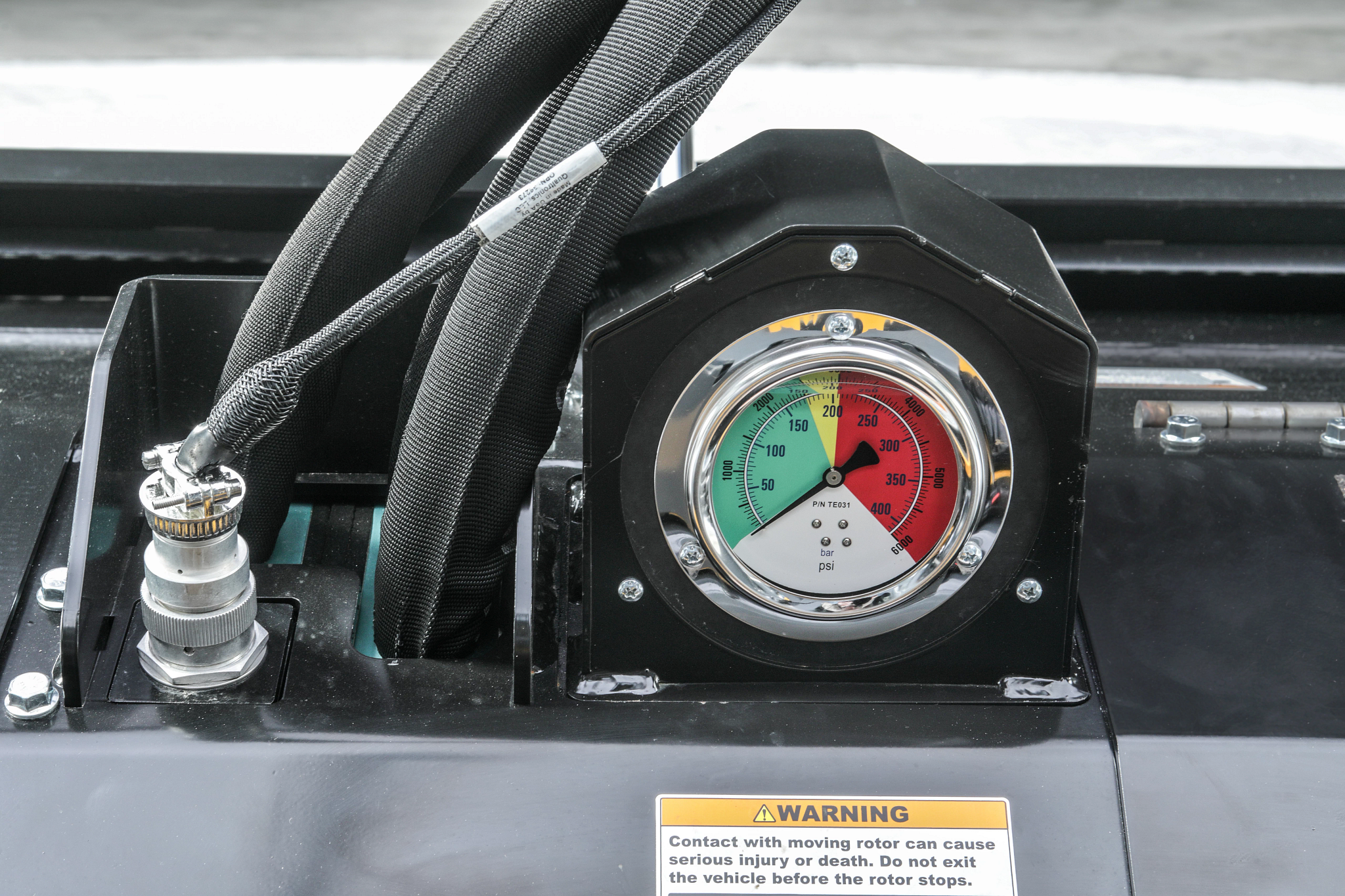 Close-up of hydraulic pressure gauge mounted, showing color-coded pressure ranges in green, yellow, and red, with braided hoses and a warning label visible on the metal housing.