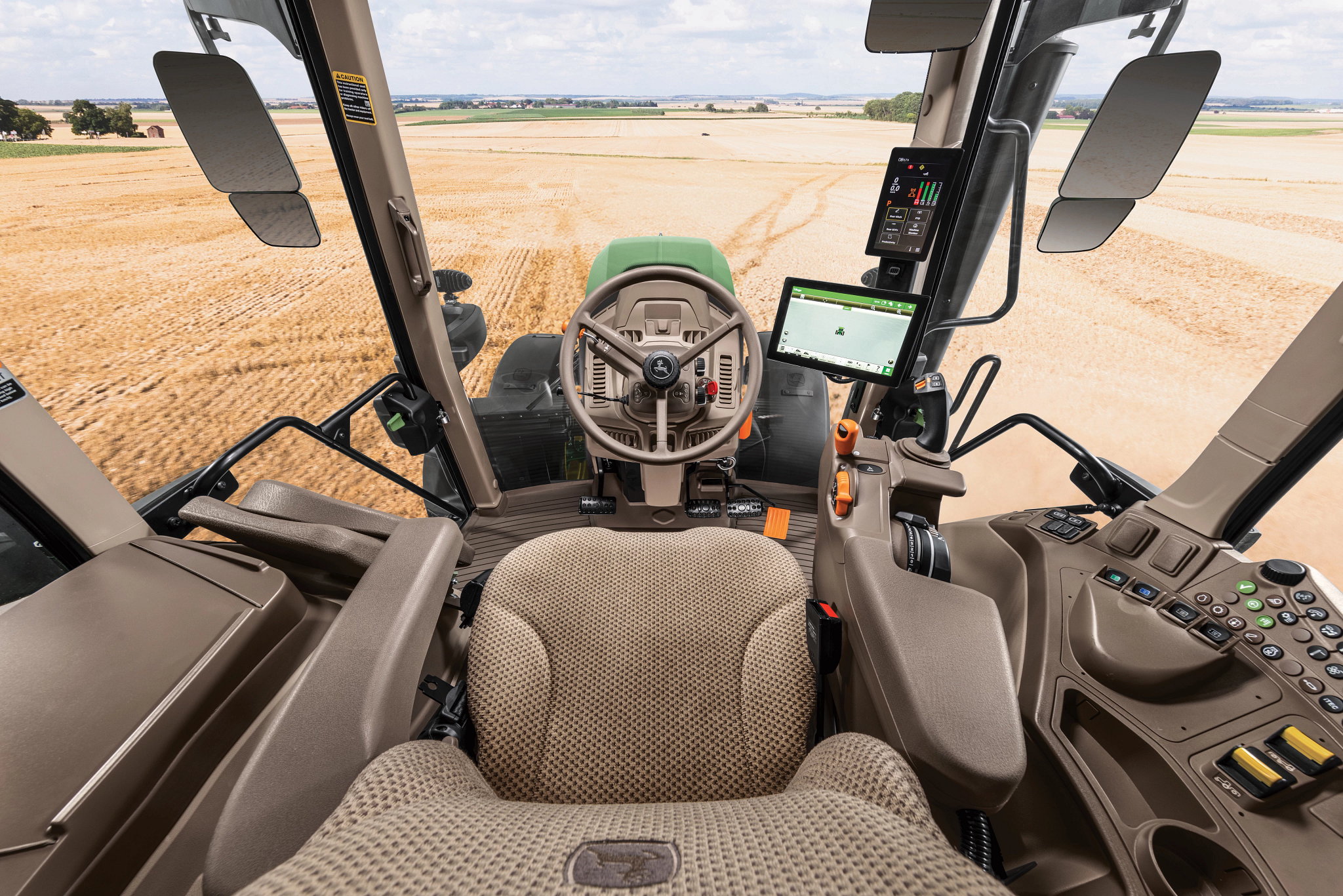 Operator’s view inside John Deere tractor cab showing fabric operator seat, steering wheel, armrest controls, touchscreen displays, and wide windshield overlooking a harvested field.