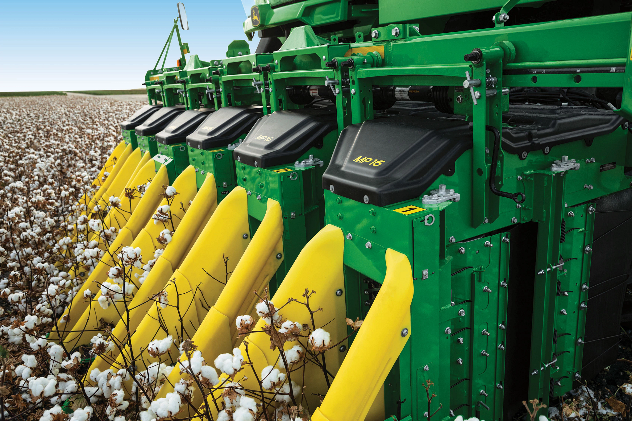 Green agriculture machinery harvesting cotton in a field, featuring multiple yellow picking heads collecting white cotton bolls under a clear sky