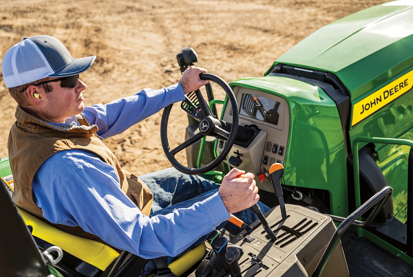 Operator seated on an open‑station John Deere tractor, holding the steering wheel and control levers; green hood with John Deere branding, instrument panel, and yellow seat visible while working on a dirt field.