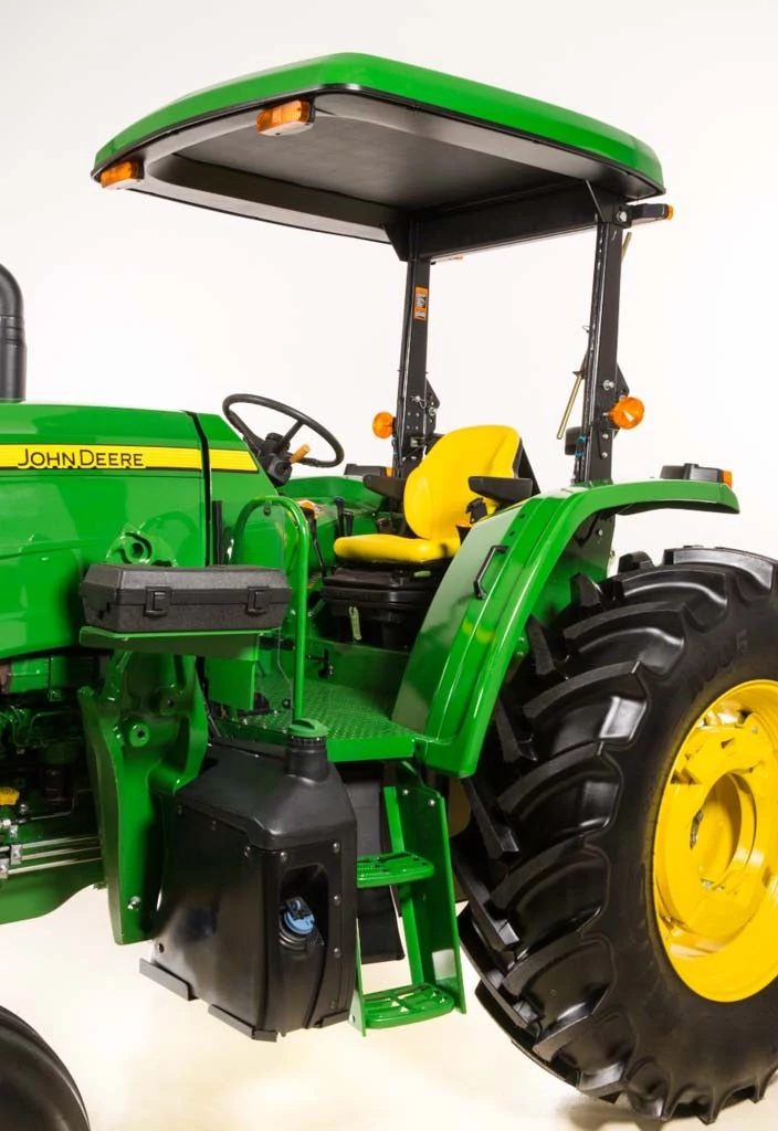 John Deere tractor operator station with open ROPS canopy, yellow seat, steering wheel, steps, and large rear tire visible, shown from the left side against a plain studio background