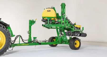 A planter folded for transport attached to a tractor hitch, featuring a large yellow tank mounted on a green frame, gauge wheels, and a row-unit section with black rubber closing wheels