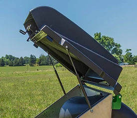 Close-up of a rear-mounted mower collection hopper with the top lid open, supported by struts, shown outdoors in a grassy field