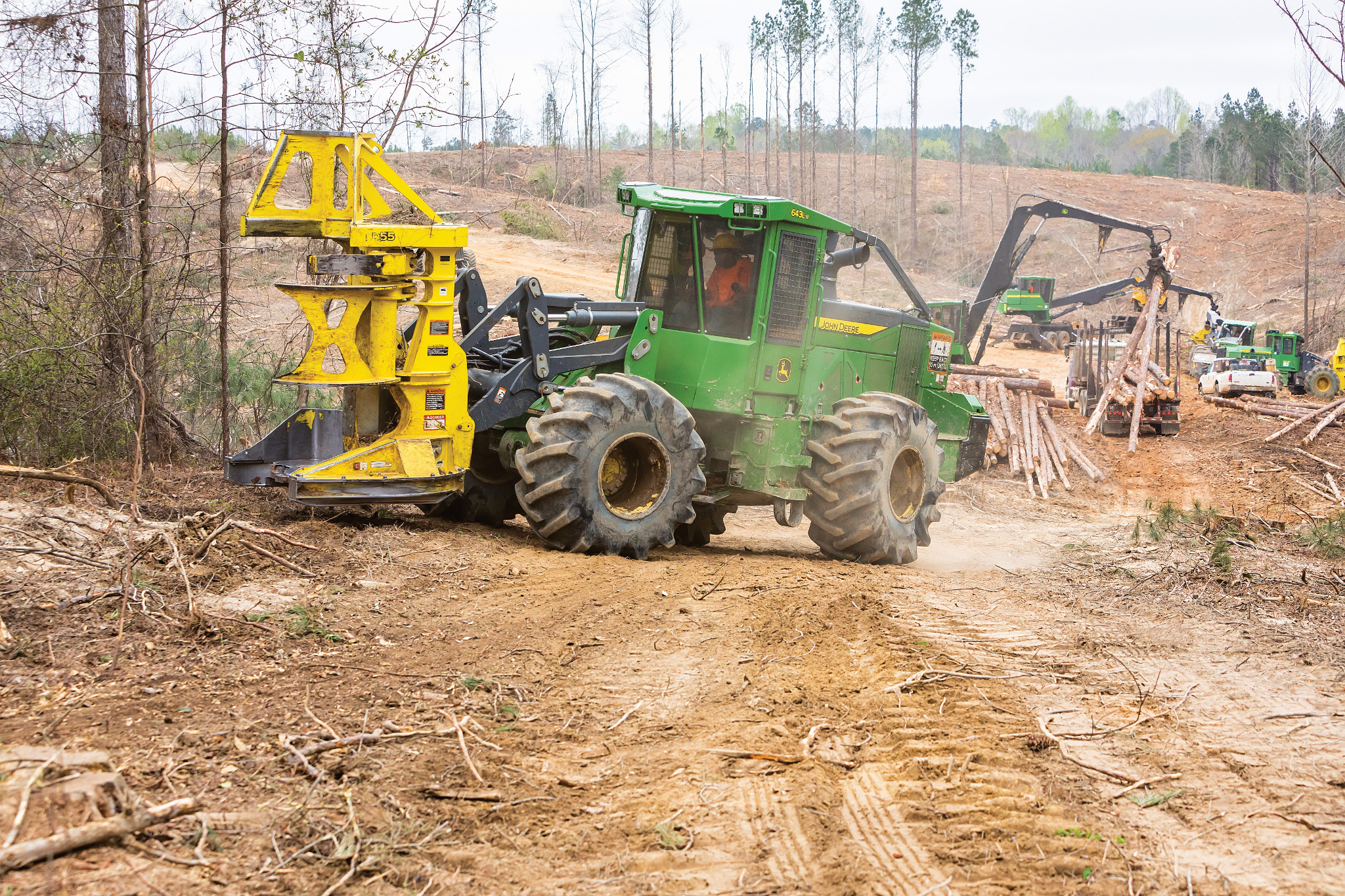 John Deere wheeled feller buncher operates in a logging site, carrying a yellow cutting head and moving along a dirt path with stacked logs and other forestry equipment visible in the background.