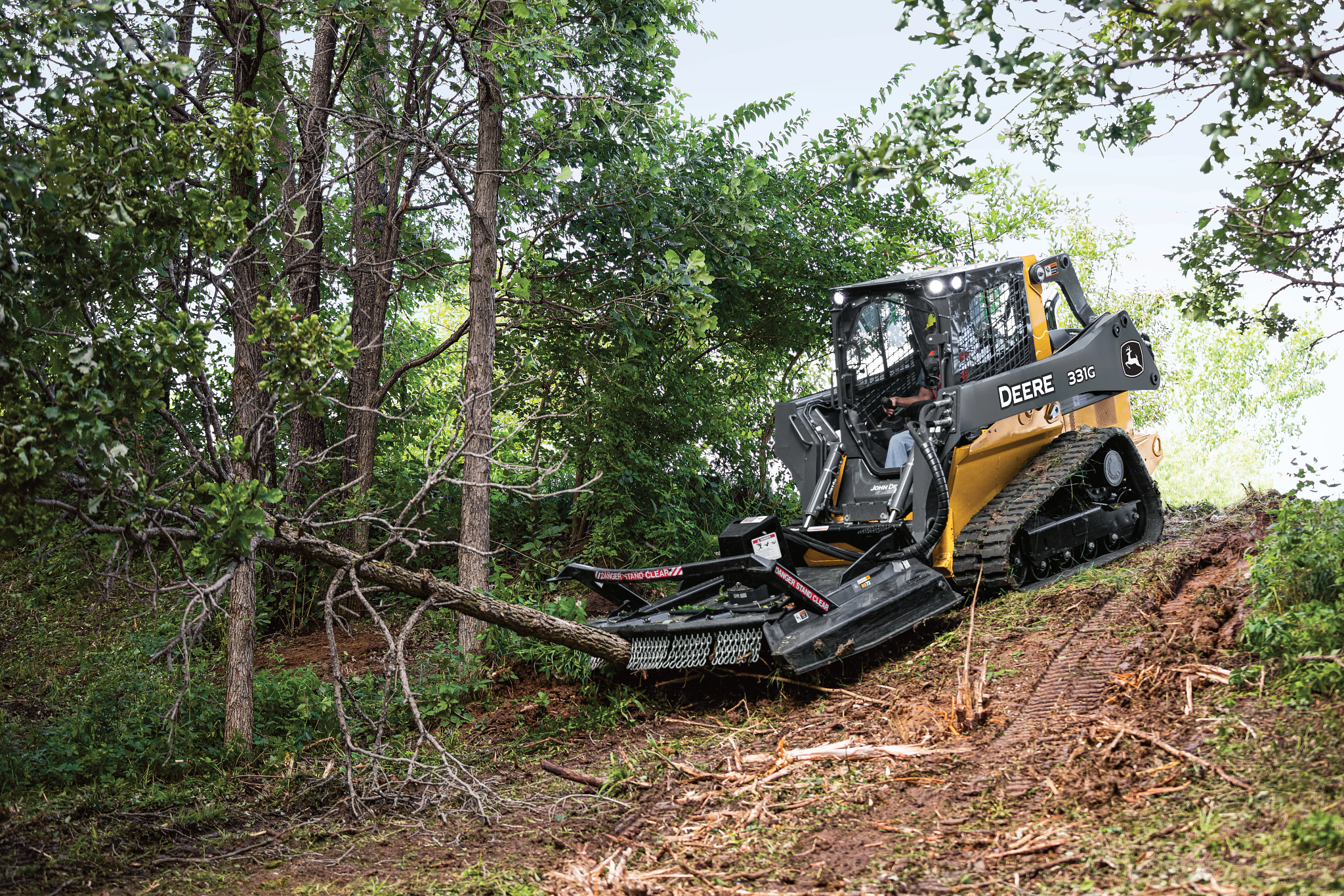Compact track loader equipped with a front rotary cutter clearing a fallen tree on a wooded slope, surrounded by trees and dense vegetation