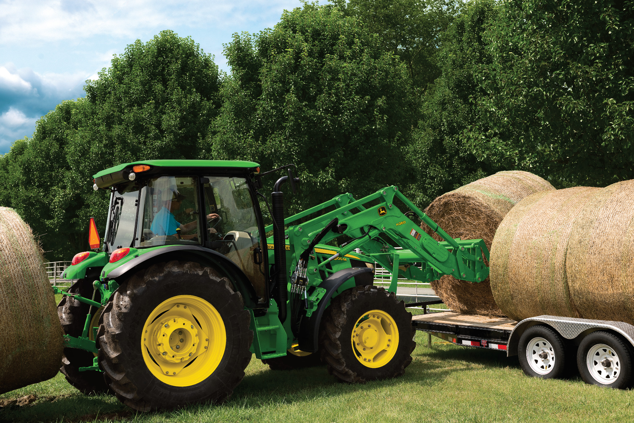 A tractor using a front loader to lift a large round hay bale from a field and place it onto a flatbed trailer, with trees in the background.