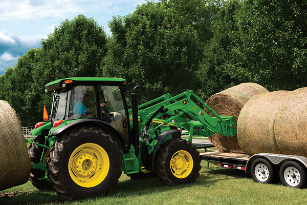 A tractor using a front loader to lift a large round hay bale from a field and place it onto a flatbed trailer, with trees in the background.