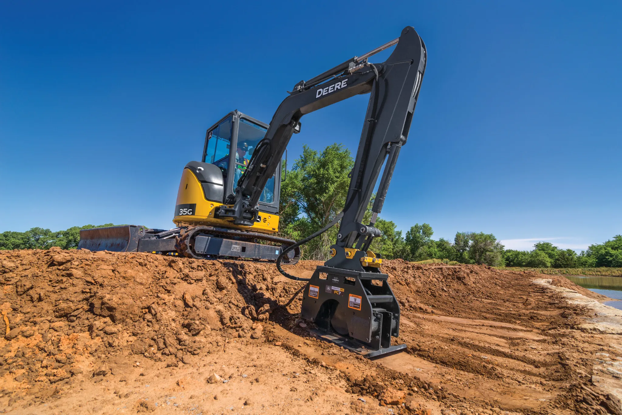 Yellow and black tracked excavator using a plate compactor attachment to smooth dirt on a large construction site, with green trees under a clear blue sky in the background