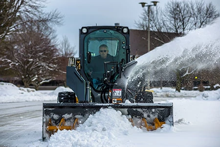 A compact loader equipped with a front snow blower clearing snow off a road, blowing snow to the side with trees and buildings in the background