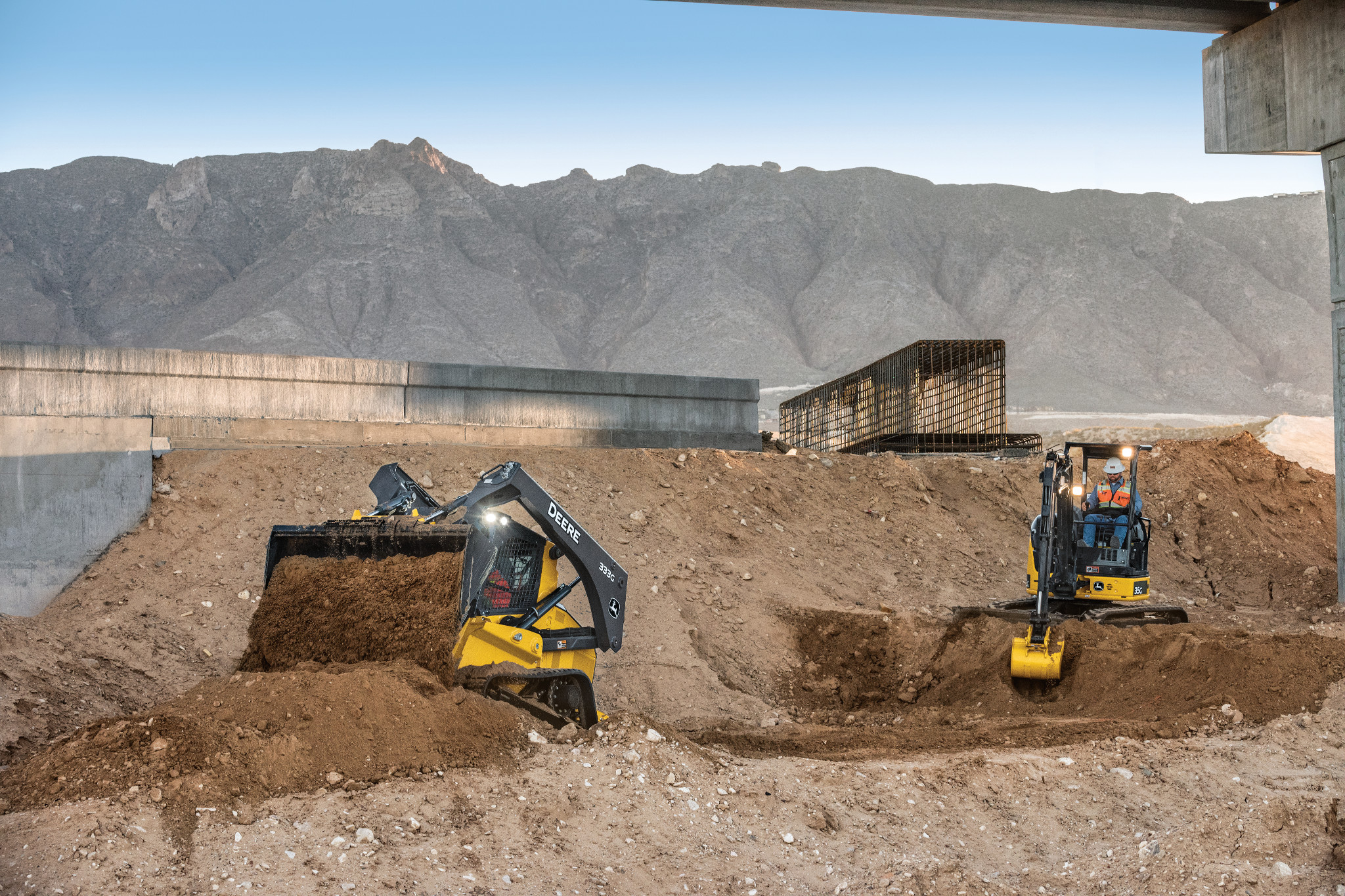 Construction site with a skid-steer loader and a compact excavator digging into dirt piles, with concrete retaining walls and a mountainous landscape in the background