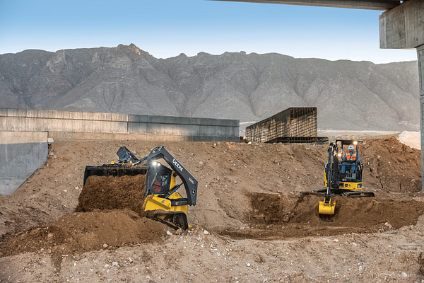 Construction site with a skid-steer loader and a compact excavator digging into dirt piles, with concrete retaining walls and a mountainous landscape in the background