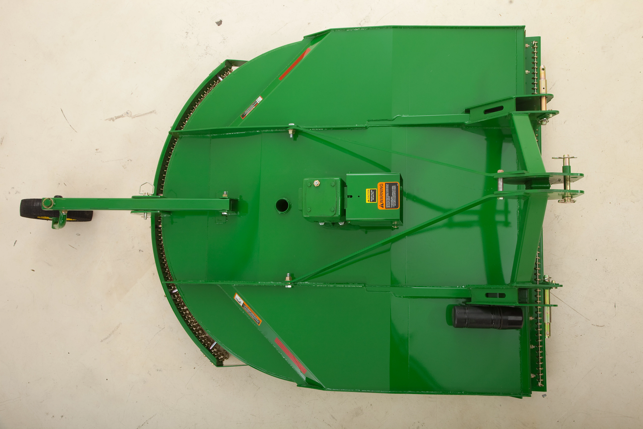 Top-down view of a green rotary cutter with a rounded deck, central gearbox, hitch arm, and protective safety labels on the surface