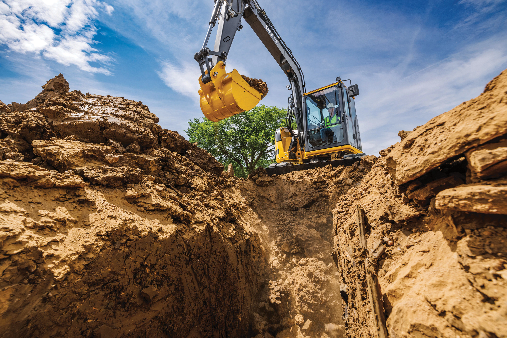 Yellow John Deere excavator with a black arm and raised bucket digging into a deep trench at a construction site