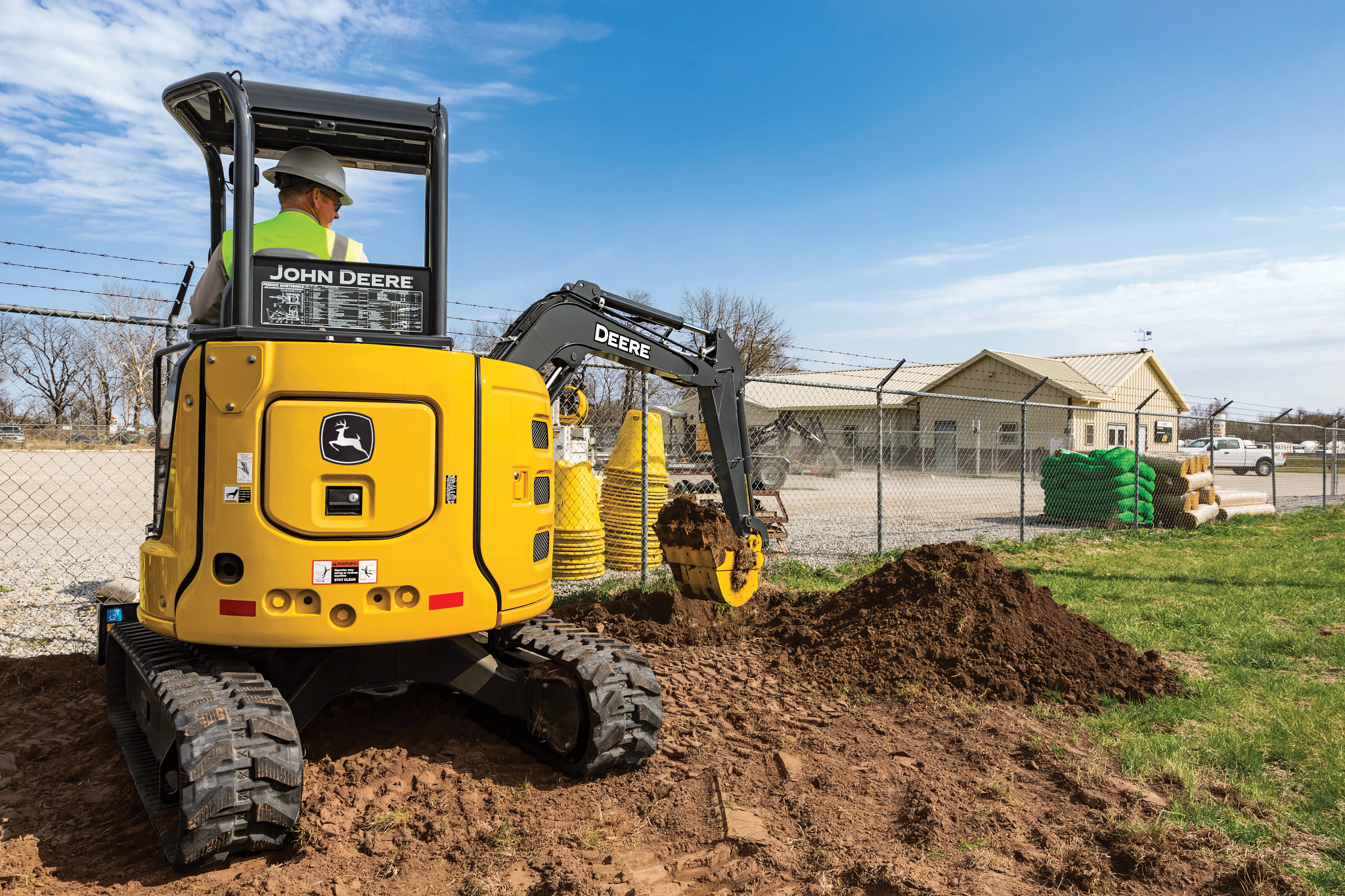 Compact John Deere excavator digging a trench beside a fence, operated by a worker in a hard hat at a grassy residential construction site