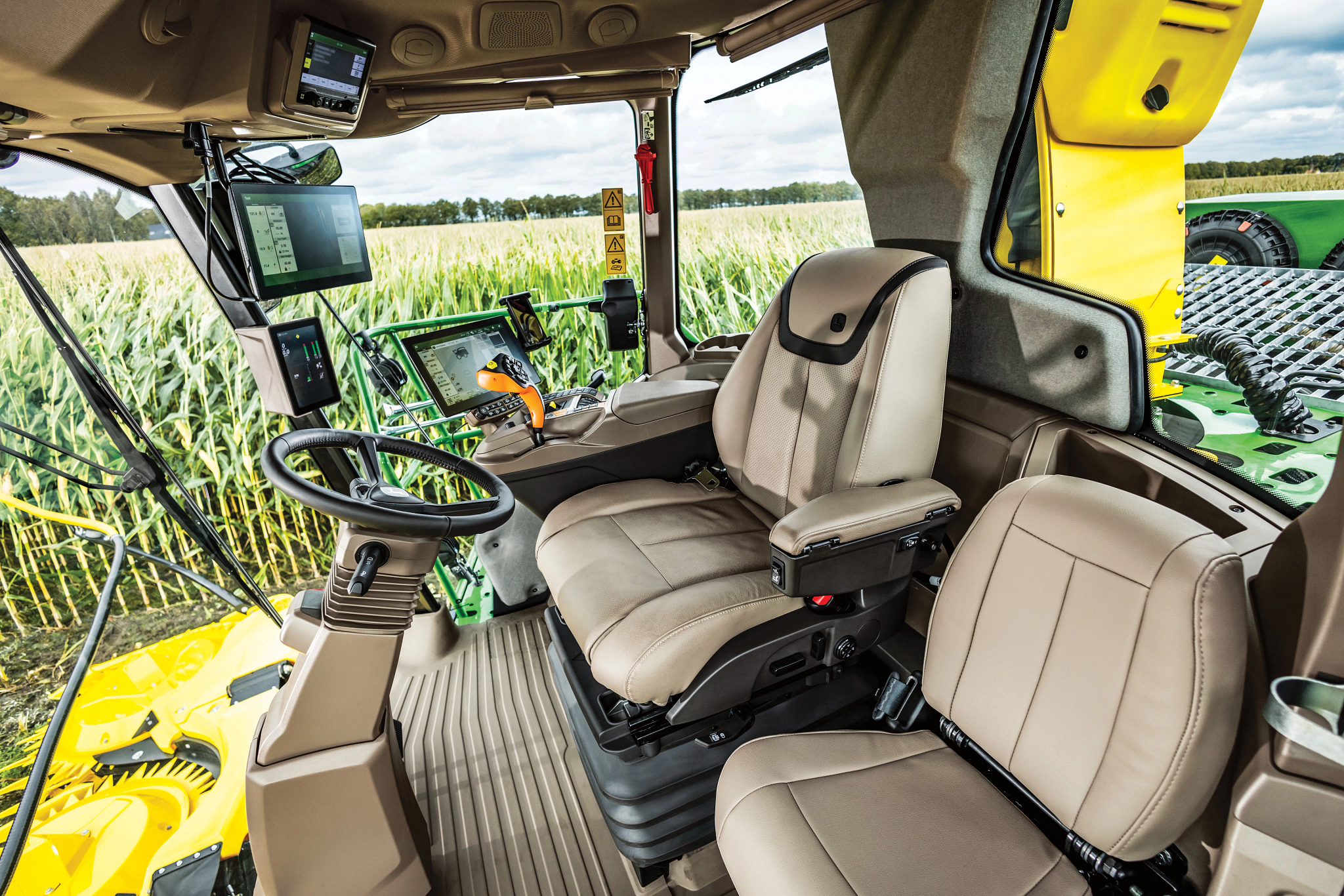 Interior of John Deere equipment cab with two beige seats, steering wheel, armrest controls, and multiple displays, viewed from the doorway with a header visible outside.