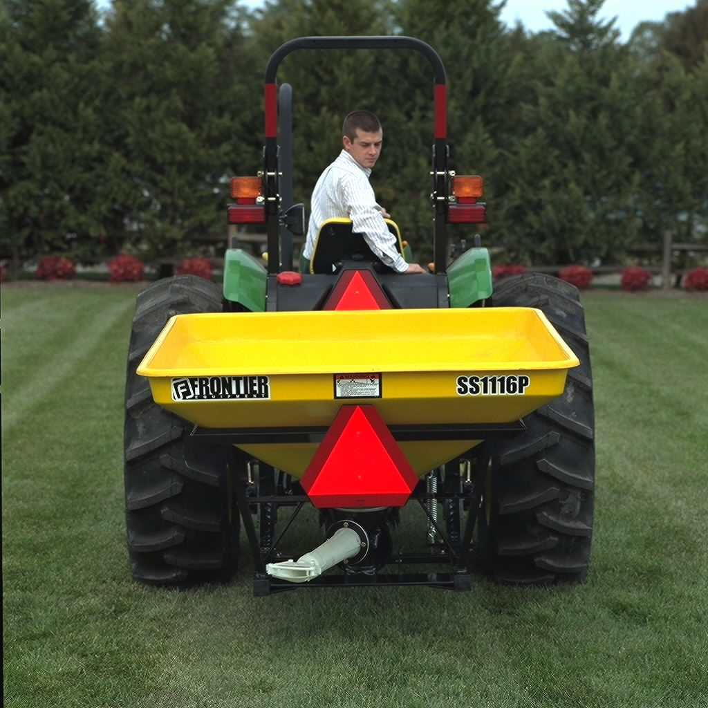 Rear view of a man driving a tractor across a lawn, equipped with a yellow Frontier SS1116P Pendular Spreader mounted on the back