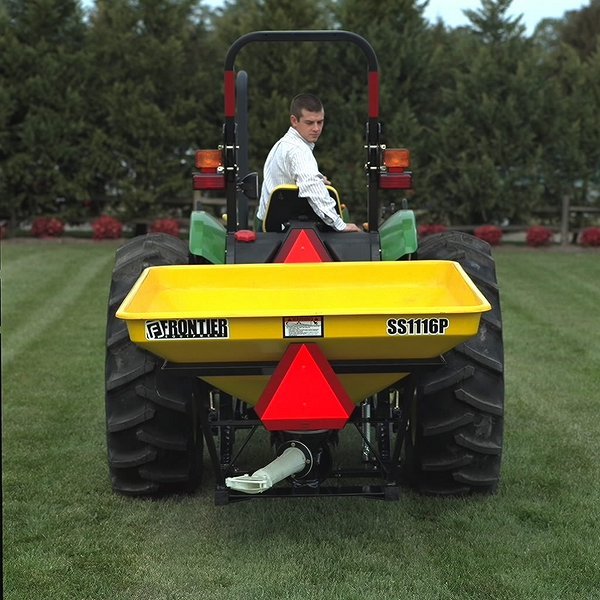 Rear view of a man driving a tractor across a lawn, equipped with a yellow Frontier SS1116P Pendular Spreader mounted on the back